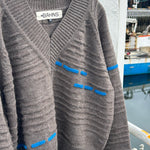 A brown ribbed knit cardigan sweater with blue contrast stitching details, hanging on a black hanger against a white wall with a blurred outdoor background.