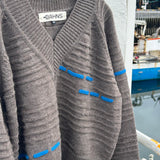 A brown ribbed knit cardigan sweater with blue contrast stitching details, hanging on a black hanger against a white wall with a blurred outdoor background.