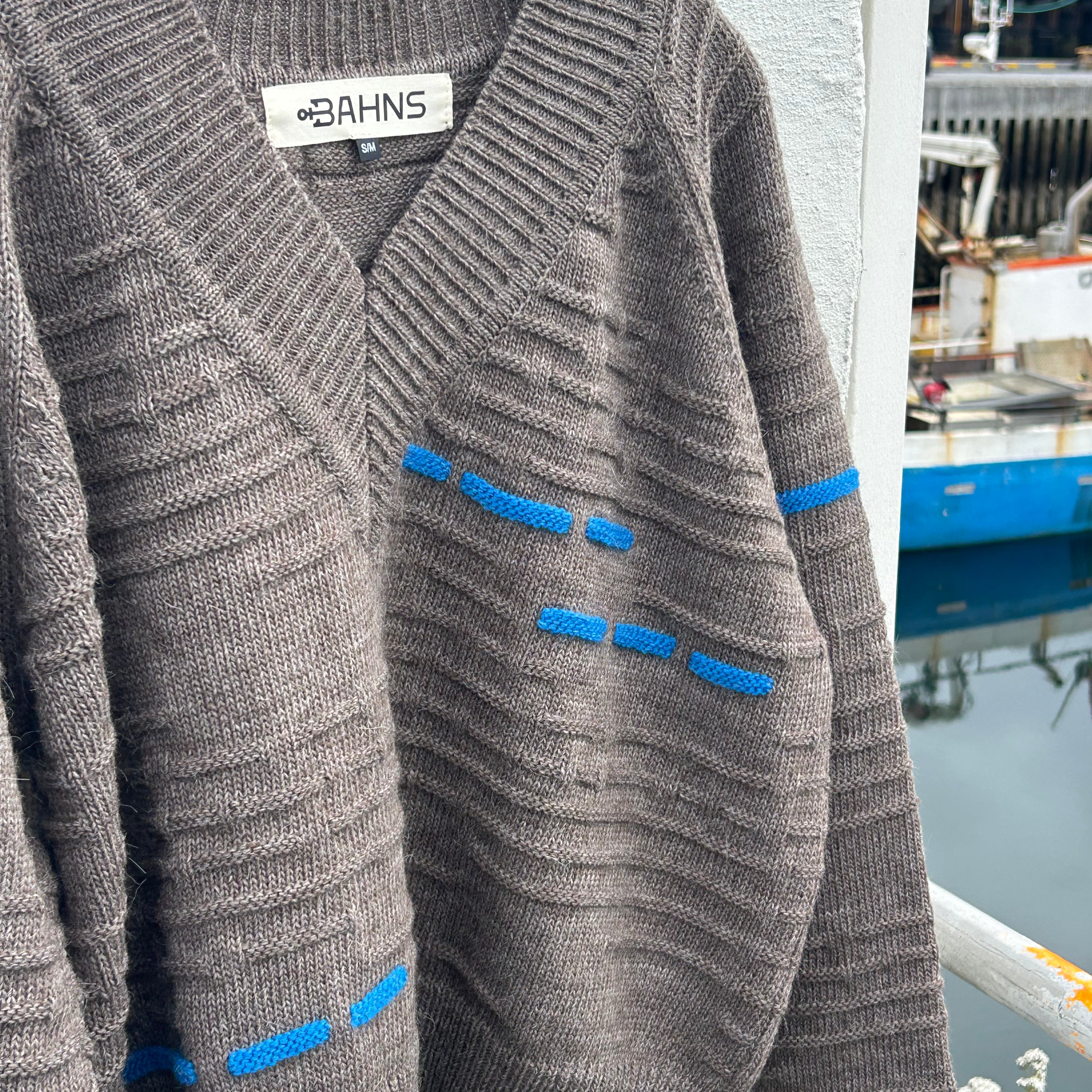 A brown ribbed knit cardigan sweater with blue contrast stitching details, hanging on a black hanger against a white wall with a blurred outdoor background.