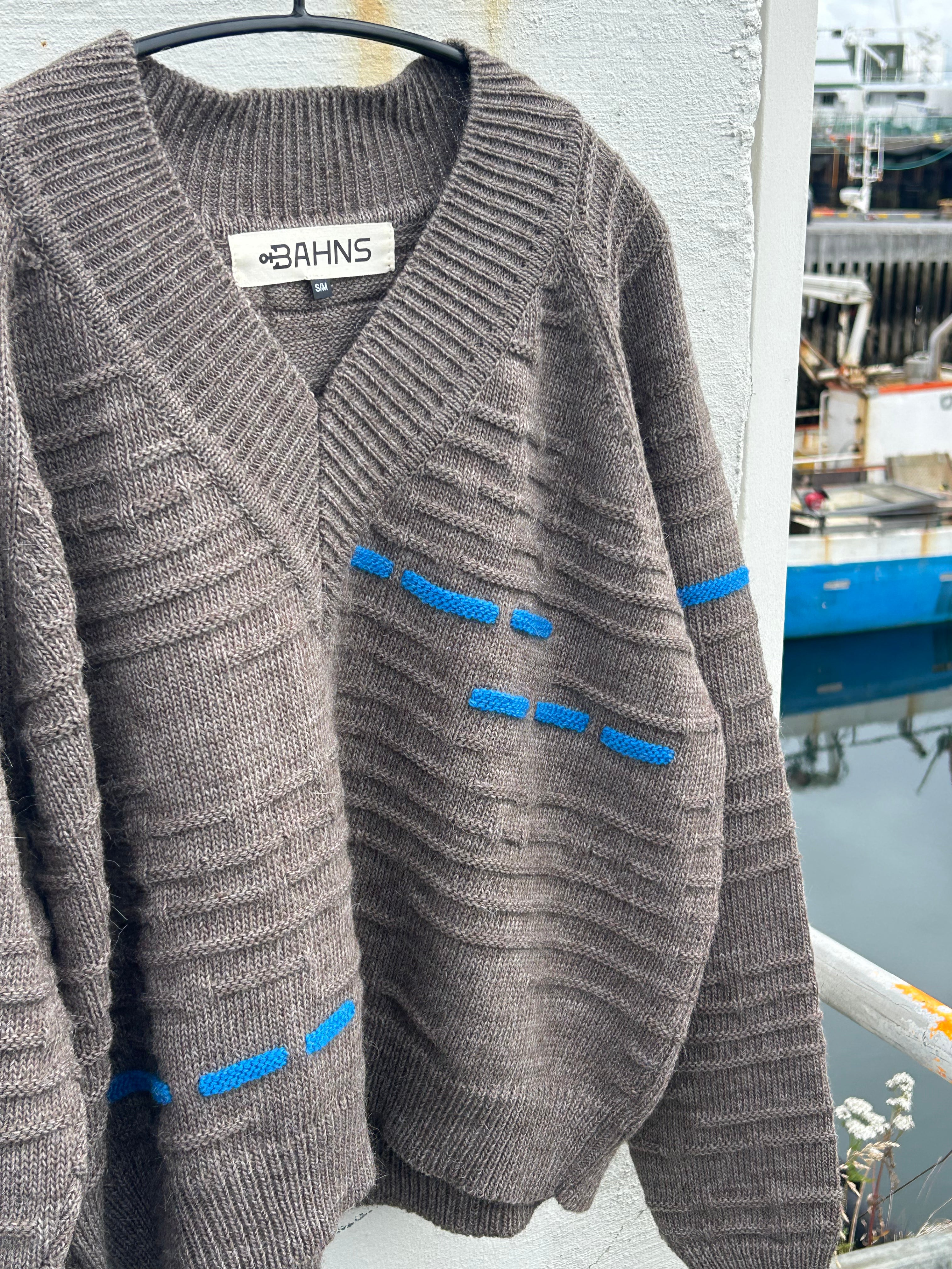 A brown ribbed knit cardigan sweater with blue contrast stitching details, hanging on a black hanger against a white wall with a blurred outdoor background.